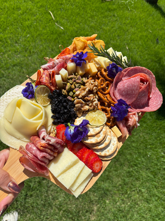 Wooden charcuterie board with various meats, cheeses, and fruits on a grassy background